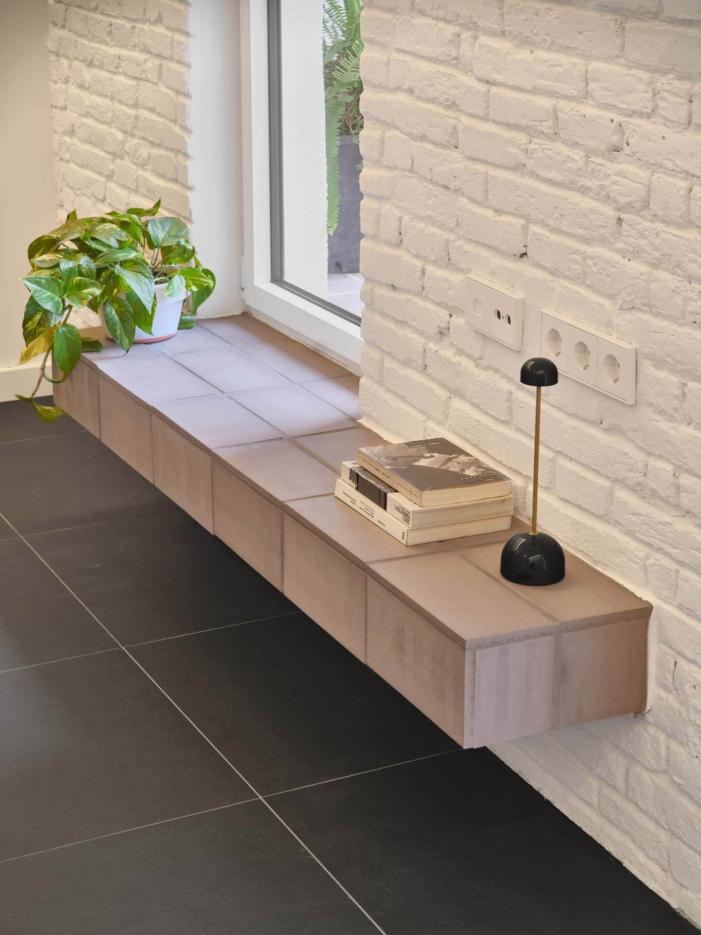 Detail from CRÜ’s “La Dolors” renovation showing a floating tiled bench against a whitewashed brick wall, with books, a small lamp, and a plant by the patio window